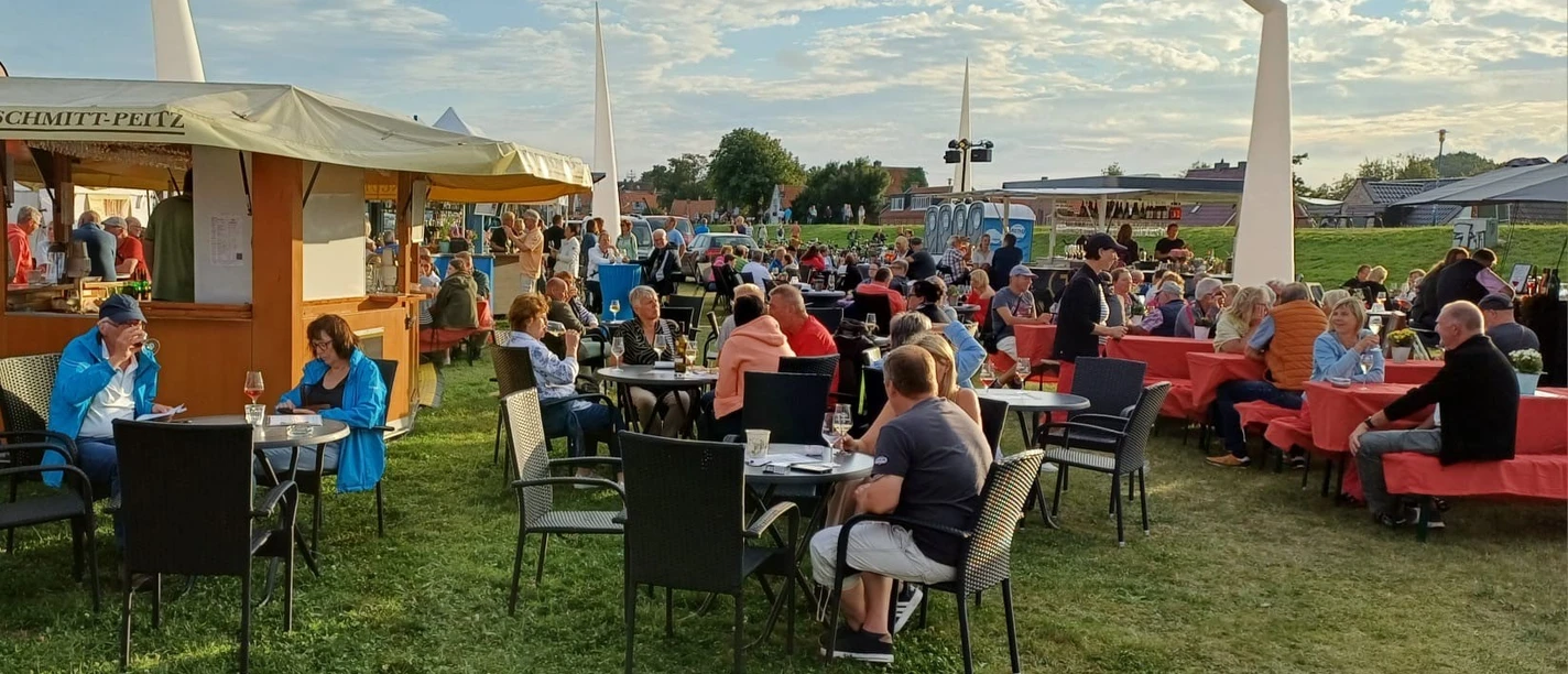 Weinfest Greetsiel Menschen sitzen an Tischen auf der Wiese am Hafen von Greetsiel und genießen Wein bei Abendsonne.