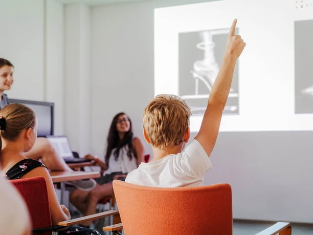 Nacht der Technik Köln Ein Junge hebt aufmerksam den Arm in einem Workshopraum, um eine Frage zu stellen.