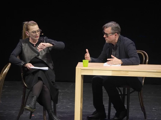 Anna Loos_Jan Josef Liefers (c) Thomas Bartilla.JPG Eine Frau und ein Mann sitzen dunkel gekleidet an einem Holztisch und reden miteinander. Die Frau schaut auf einen Papierblock, während der Mann sie anschaut.