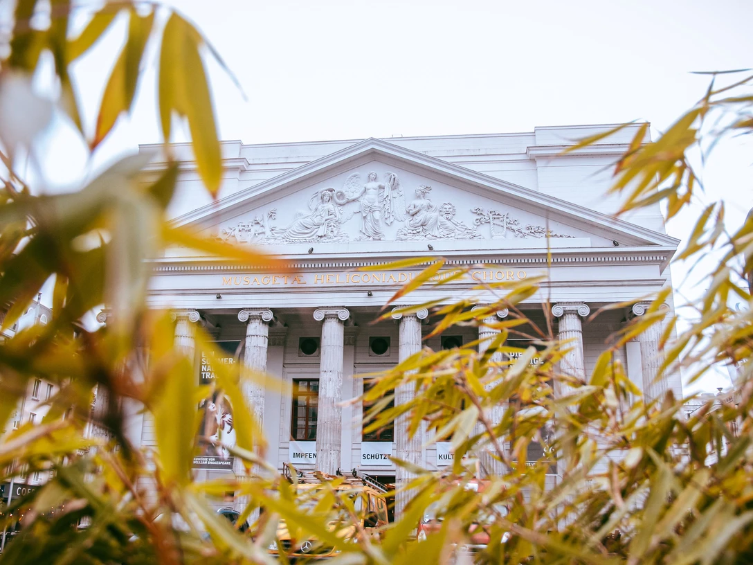 Hinter herbstlich grünen und orangenen Blättern kommt die kunstvolle Fassade des Theaters Aachen mit seinen Säulen hervor.