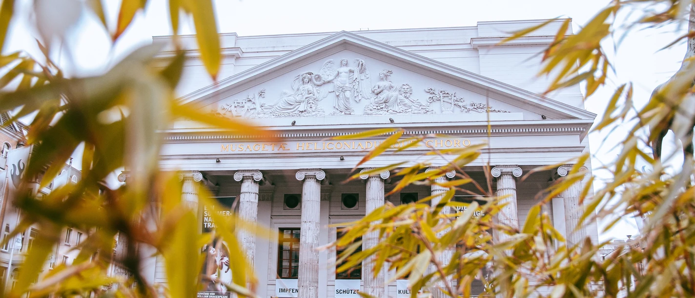 Hinter herbstlich grünen und orangenen Blättern kommt die kunstvolle Fassade des Theaters Aachen mit seinen Säulen hervor.