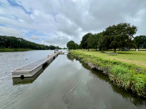Schiffsanleger Heisterholz Schiffsanleger neben ruhigem Fluss, umgeben von grünen Wiesen und Bäumen unter bewölktem Himmel.