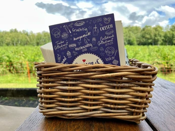 Eine geflochtene Korbkiste auf einem Holztisch enthält eine Speisekarte vor grüner Landschaft.
