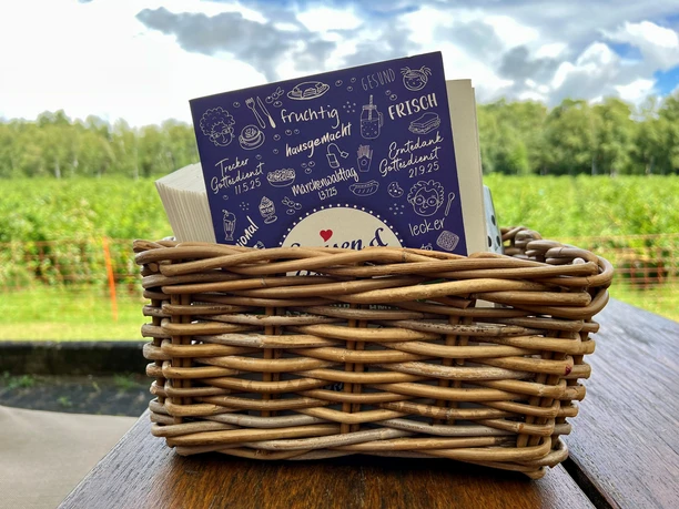 Eine geflochtene Korbkiste auf einem Holztisch enthält eine Speisekarte vor grüner Landschaft.