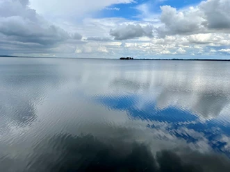 Weitblick auf ruhigen See mit spiegelnden Wolken und fernen kleinen Inseln unter wechselhaftem Himmel.