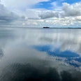 Weitblick auf ruhigen See mit spiegelnden Wolken und fernen kleinen Inseln unter wechselhaftem Himmel.
