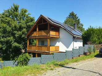 Aussenansicht Ein zweistöckiges Haus mit Holzverkleidung und Solarpanelen, umgeben von Zaun und Bäumen an sonnigem Tag.