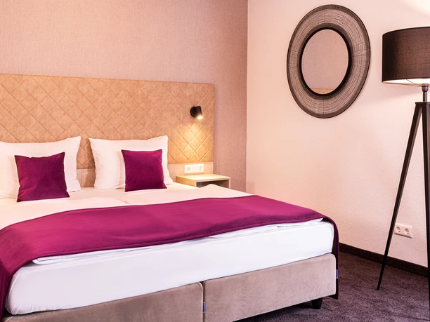 Victor’s Residenz-Hotel Gummersbach Modern eingerichtetes Hotelzimmer mit Doppelbett, beigen Kopfteil, lila Akzenten und Stehleuchte.
