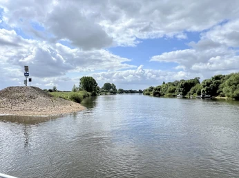 Flusslauf mit bewaldeten Ufern unter einem Himmel mit weißen Wolken und blauem Himmel im Hintergrund.