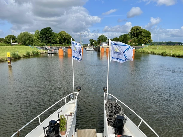 Auf einem Fluss fährt ein Ausflugsschiff auf eine geöffnete Klappbrücke zu, umgeben von grüner Landschaft.