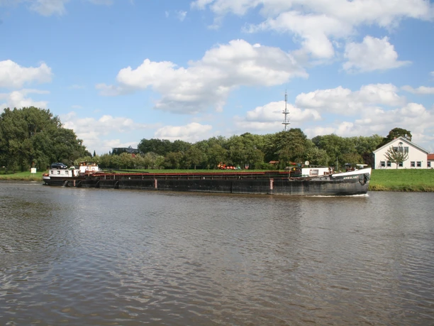 A large freighter sails on a calm river near Nienburg, surrounded by green trees and a clear sky.