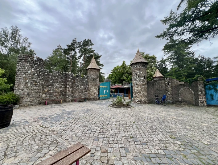 Ein steinernes Burgtor mit drei Türmen vor einem bewölkten Himmel, umgeben von Bäumen und Gehweg.A stone castle gate with three towers against a cloudy sky, surrounded by trees and sidewalk.En borgport af sten med tre tårne mod en overskyet himmel, omgivet af træer og fortov.Een stenen kasteelpoort met drie torens tegen een bewolkte hemel, omringd door bomen en bestrating.