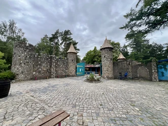 Magic Park Verden Ein steinernes Burgtor mit drei Türmen vor einem bewölkten Himmel, umgeben von Bäumen und Gehweg.