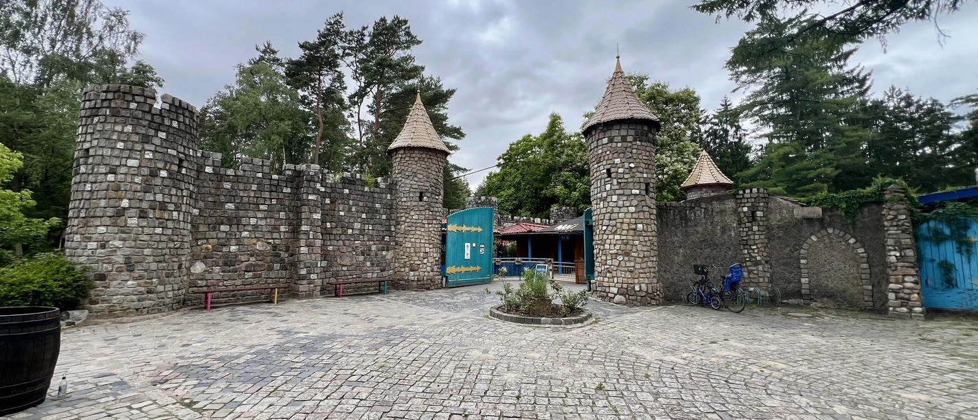 Magic Park Verden A stone castle gate with three towers against a cloudy sky, surrounded by trees and sidewalk.