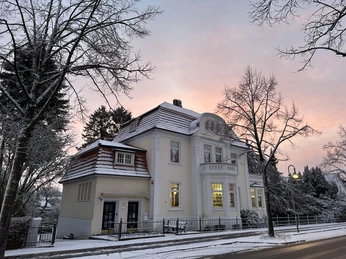 2025-01-04_Schnee (9).jpg Ein elegantes, gelb gestrichenes Gebäude im Schnee bei Sonnenaufgang, umgeben von kahlen Bäumen.