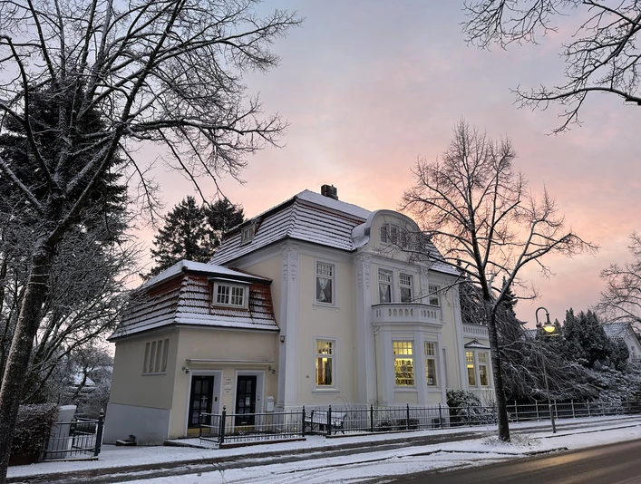 2025-01-04_Schnee (9).jpg Ein elegantes, gelb gestrichenes Gebäude im Schnee bei Sonnenaufgang, umgeben von kahlen Bäumen.