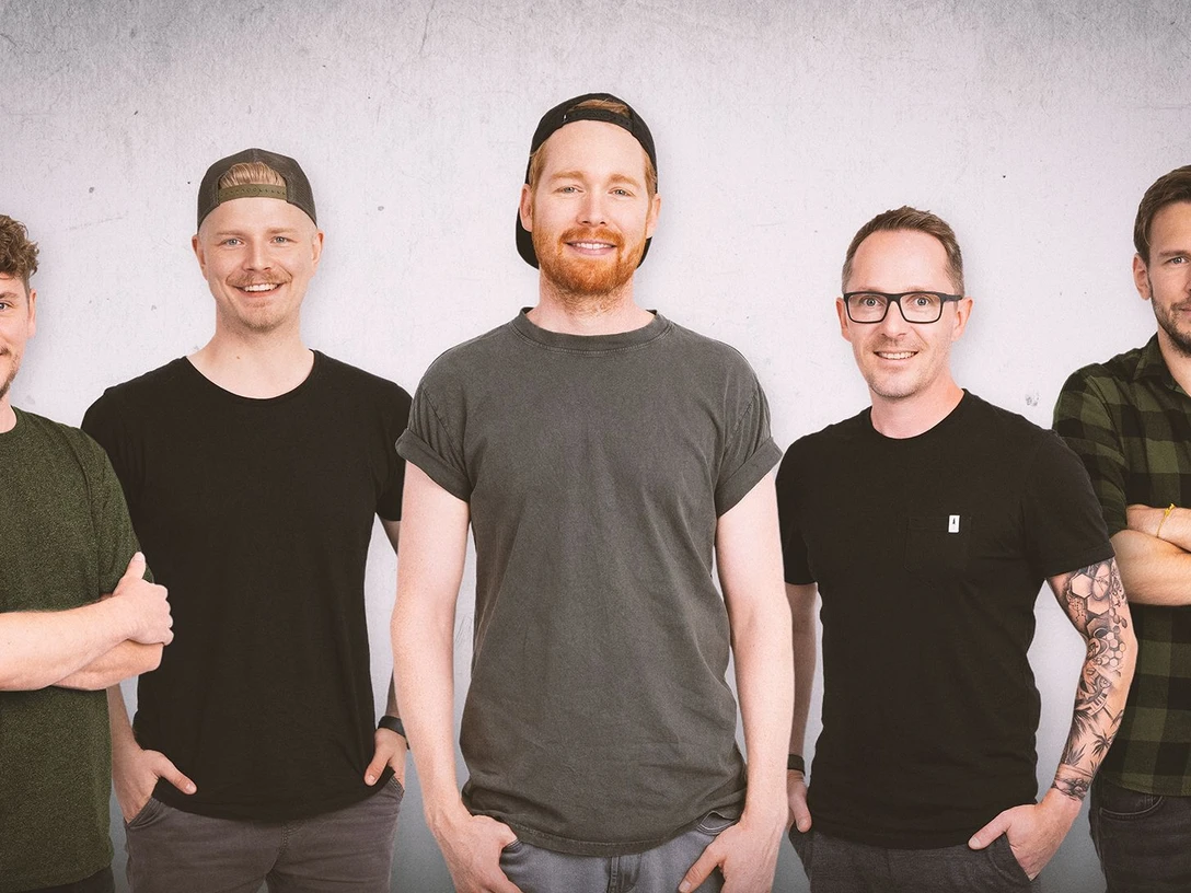 Fünf junge Männer in dunkler Kleidung stehen nebeneinander vor weißem Hintergrund und lächeln in die Kamera.