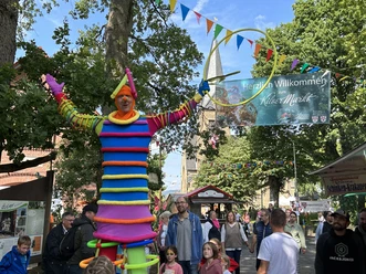 Ein bunt gekleideter Stelzenläufer auf einem belebten Stadtfest mit neugierigen Besuchern.