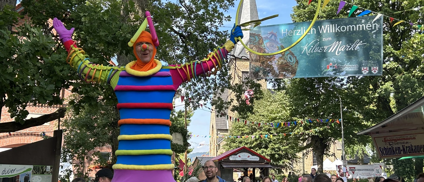 Ein bunt gekleideter Stelzenläufer auf einem belebten Stadtfest mit neugierigen Besuchern.