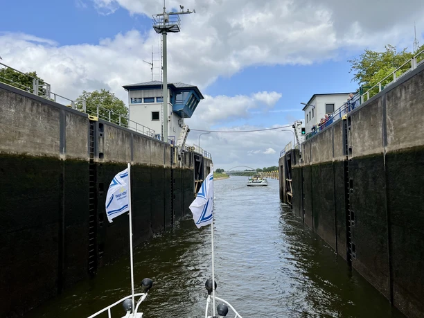 Schleuse in Betrieb mit geöffneten Toren, dahinter Wasserweg durch grüne Landschaft, bedeckter Himmel.