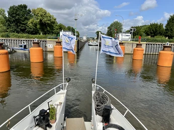 Zwei Boote fahren auf eine geöffnete Schleuse zu, flankiert von leuchtend orangen Pollern.