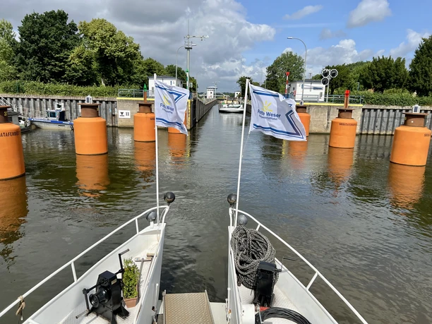 Zwei Boote fahren auf eine geöffnete Schleuse zu, flankiert von leuchtend orangen Pollern.