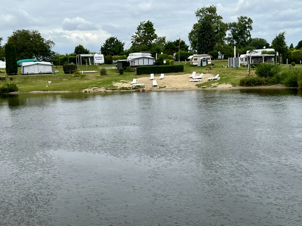 Ein ruhiger Campingplatz am Wasser mit Wohnwagen, Rasenfläche und mehreren Liegestühlen am Ufer.