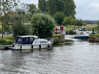 Motorboote liegen am Steg eines ruhigen Flussufers, umgeben von üppigem Grün und bewölktem Himmel.