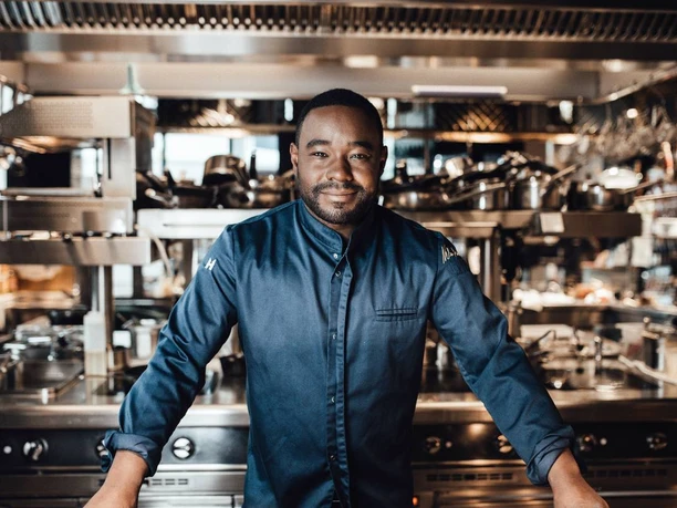 Nelson Müller Ein Koch in dunkelblauer Uniform steht lächelnd in einer modernen, professionellen Restaurantküche.