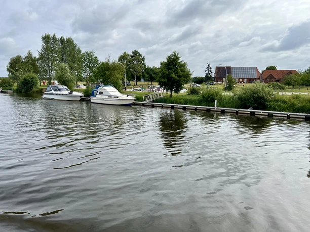 Boote liegen am ruhigen Ufer eines Flusses, umgeben von Bäumen und einer ländlichen Landschaft.