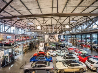 MS Hangar A large, modern hangar with an impressive collection of vehicles. Various sports cars in bright colors, such as red, blue and white, are displayed on the hangar floor. Above left there is a gallery level with further exhibits. The roof construction is made of steel and gives the room an industrial charm. Light emanates from several luminaires and creates a bright, inviting atmosphere.