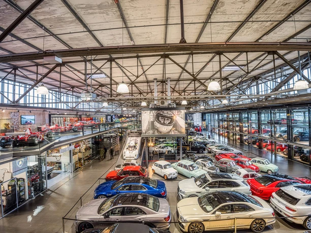 MS Hangar A large, modern hangar with an impressive collection of vehicles. Various sports cars in bright colors, such as red, blue and white, are displayed on the hangar floor. Above left there is a gallery level with further exhibits. The roof construction is made of steel and gives the room an industrial charm. Light emanates from several luminaires and creates a bright, inviting atmosphere.