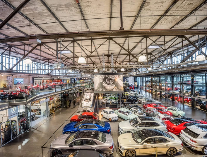 MS Hangar Ein großer, modernen Hangars mit einer beeindruckenden Sammlung von Fahrzeugen. Verschiedene Sportwagen in leuchtenden Farben, wie Rot, Blau und Weiß, sind auf dem Hallenboden ausgestellt. Oben links gibt es eine Galerieebene mit weiteren Exponaten. Die Dachkonstruktion ist aus Stahl und verleiht dem Raum einen industriellen Charme. Licht geht von mehreren Leuchten ab und sorgt für eine helle, einladende Atmosphäre.A large, modern hangar with an impressive collection of vehicles. Various sports cars in bright colors, such as red, blue and white, are displayed on the hangar floor. Above left there is a gallery level with further exhibits. The roof construction is made of steel and gives the room an industrial charm. Light emanates from several luminaires and creates a bright, inviting atmosphere.