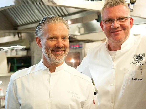 Brüder Wilbrand Zwei lächelnde Köche in weißer Kochkleidung stehen in einer professionellen Restaurantküche.