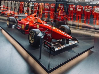 Ferrari Michael Schumacher Ein roter Ferrari-Rennwagen steht in einer gläsernen Vitrine, umgeben von Rennanzügen.A red Ferrari racing car stands in a glass display case, surrounded by racing suits.