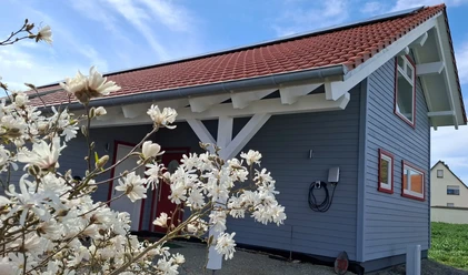 Finkennest.jpg Blaues Holzhaus mit rotem Dach, umgeben von blühendem Magnolienstrauch unter klarem Himmel.