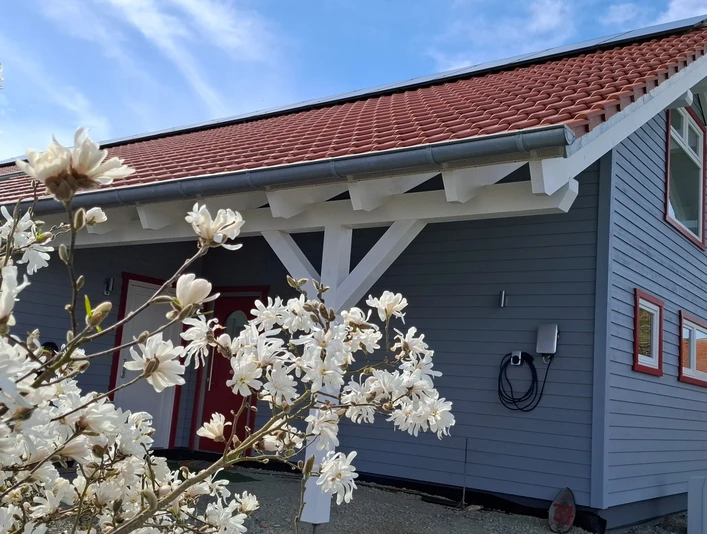 Finkennest.jpg Blaues Holzhaus mit rotem Dach, umgeben von blühendem Magnolienstrauch unter klarem Himmel.