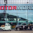 Motorworld Cologne Ein schwarzer Pickup-Truck mit einem Motorrad auf der Ladefläche und ein weißer Caddy stehen vor der gläsernen Fassade der Motorworld Köln, auf der das rot-graue Motorworld-Logo prangt.A black pickup truck with a motorcycle on the back and a white Caddy are parked in front of the glass façade of Motorworld Köln, emblazoned with the red and gray Motorworld logo.