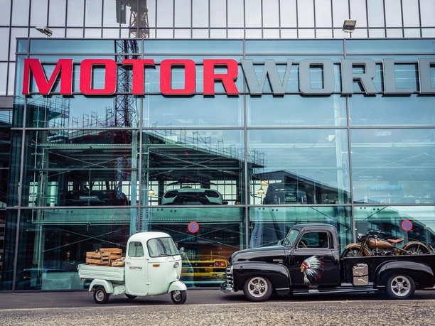 MOTORWORLD Köln Rheinland Ein schwarzer Pickup-Truck mit einem Motorrad auf der Ladefläche und ein weißer Caddy stehen vor der gläsernen Fassade der Motorworld Köln, auf der das rot-graue Motorworld-Logo prangt.