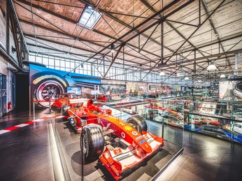 F1 Ferrari Michael Schumacher Motorworld Cologne Ein roter Formel-1-Ferrari in einer lichtdurchfluteten Ausstellungshalle. Die Umgebung ist modern gestaltet, mit einer hohen Decke und großen Fenstern, durch die viel Tageslicht fällt. Im Hintergrund sind weitere Exponate zu erkennen, die das Motorsporterlebnis ergänzen. Der Ferrari steht im Vordergrund auf einem Podest, begleitet von einer Atmosphäre der Geschwindigkeit und Eleganz.A red Formula 1 Ferrari in a light-flooded exhibition hall. The surroundings are modern, with a high ceiling and large windows that let in plenty of natural light. Other exhibits can be seen in the background, complementing the motorsport experience. The Ferrari stands on a pedestal in the foreground, accompanied by an atmosphere of speed and elegance.