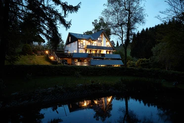 Außenansicht in der Dämmerung Ein idyllisches Haus am Waldrand beleuchtet die Abenddämmerung, spiegelt sich im ruhigen Teich.