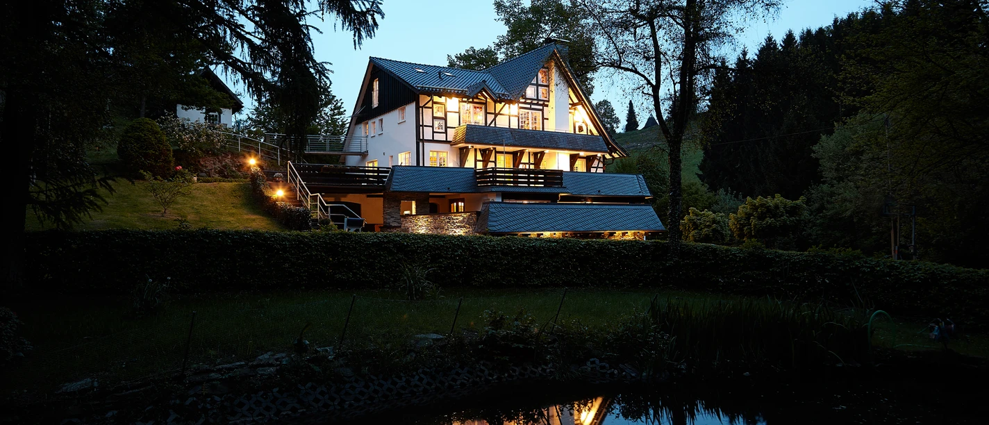 Außenansicht in der Dämmerung Ein idyllisches Haus am Waldrand beleuchtet die Abenddämmerung, spiegelt sich im ruhigen Teich.