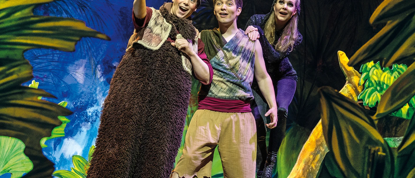 Dschungelbuch - das Musical_Pressebild 01_quer.jpg Drei Darsteller in Kostümen bewegen sich fröhlich auf einer tropischen Dschungel-Bühne.
