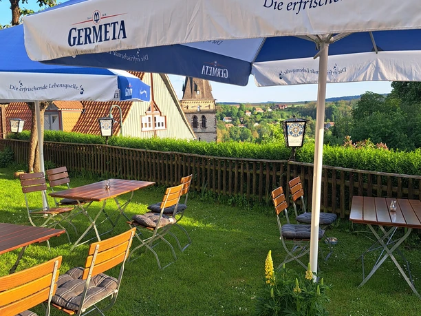 20240509_191522.jpg Biergarten mit Holztischen und Stühlen auf grüner Wiese, überdacht von blauen Sonnenschirmen, Kirchturm im Hintergrund.