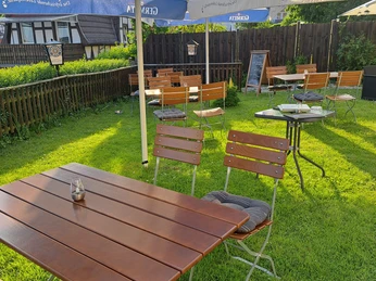 20240509_191403.jpg Ein Biergarten mit Holztischen und blauen Sonnenschirmen auf einer gepflegten Rasenfläche.