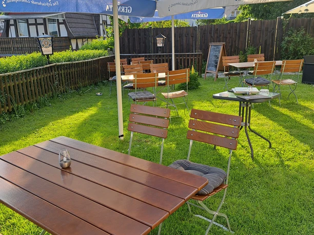 20240509_191403.jpg Ein Biergarten mit Holztischen und blauen Sonnenschirmen auf einer gepflegten Rasenfläche.