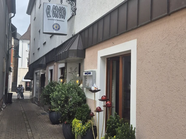 1000087001.jpg Schmale gepflasterte Gasse mit dekorierten Blumenkübeln und einer Wandbeschilderung eines Geschirrgroßhandels.