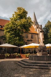 IMG_5376.JPG Außenbereich einer Gaststätte mit weißen Sonnenschirmen, umgeben von Bäumen und einer Statue im Vordergrund.