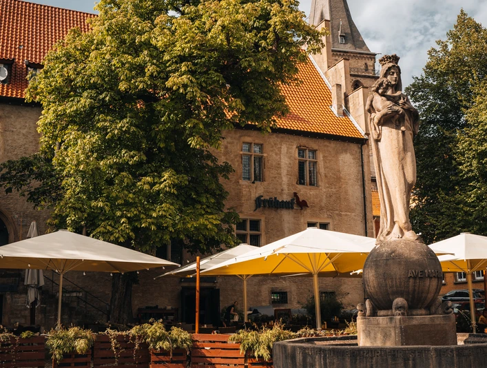 IMG_5376.JPG Außenbereich einer Gaststätte mit weißen Sonnenschirmen, umgeben von Bäumen und einer Statue im Vordergrund.
