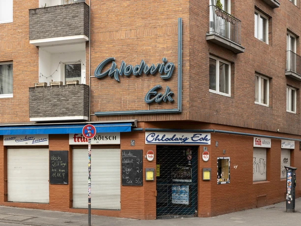 Chlodwig Eck Das Bild zeigt die Backsteinfassade der Eckkneipe "Chlodwig Eck" in Köln mit blauer Neonaufschrift.